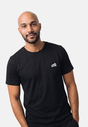Uomo calvo sorridente con barba che indossa una t-shirt nera a maniche corte con un piccolo logo bianco "WORKS" sul petto, in piedi davanti a uno sfondo semplice.