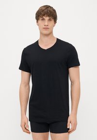 Μαύρο κοντομάνικο μπλουζάκι V-ντεκολτέ φτιαγμένο από μαλακό ύφασμα, με χαλαρή εφαρμογή και απλό σχέδιο, φορεμένο με στενές μαύρες βερμούδες.