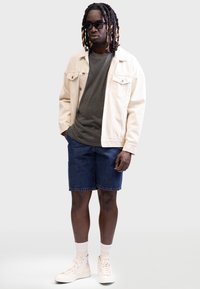 Denim shorts, grey t-shirt, cream oversized jacket, and white high-top sneakers with accents. Worn with dark sunglasses.