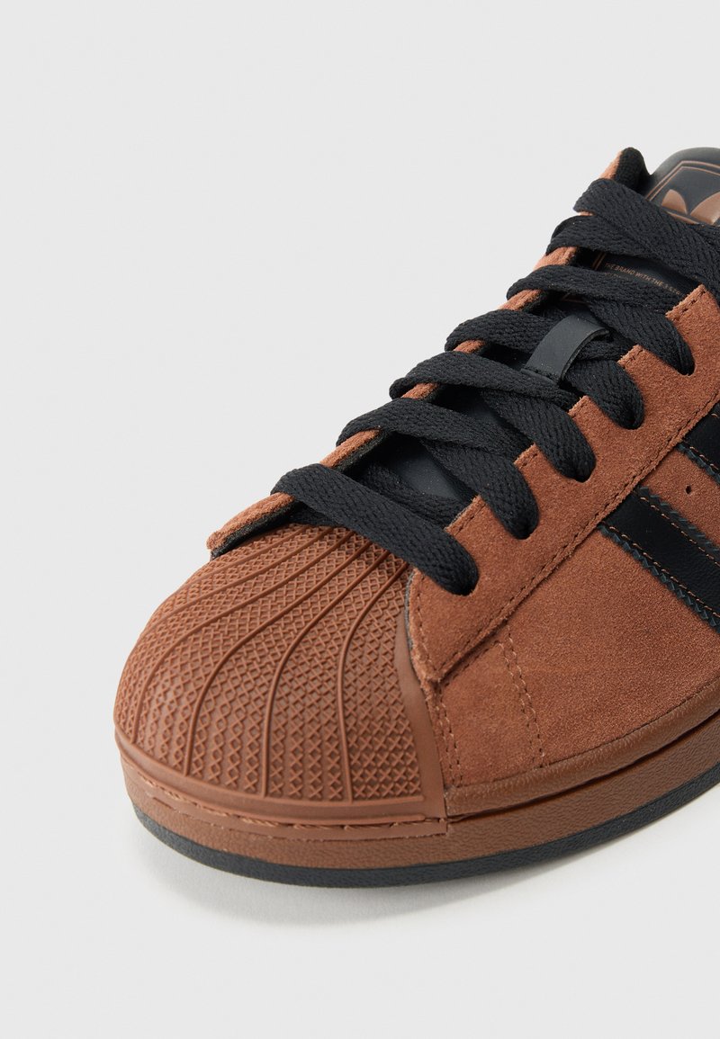 Brauner Wildleder-Sneaker mit schwarzen Schnürsenkeln und Streifen, mit einer strukturierten Gummischale an der Spitze und einer schwarzen Sohle auf weißem Hintergrund.