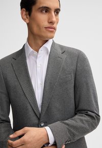 Blazer grigio realizzato in tessuto testurizzato, con revers a punta e chiusura a un bottone, abbinato a una camicia bianca.