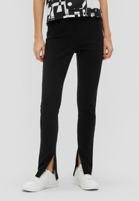 Pantalons noirs extensibles avec fentes sur les côtés, présentant une texture lisse et une coupe slim. Portés avec des baskets blanches et un haut à motif monochrome.