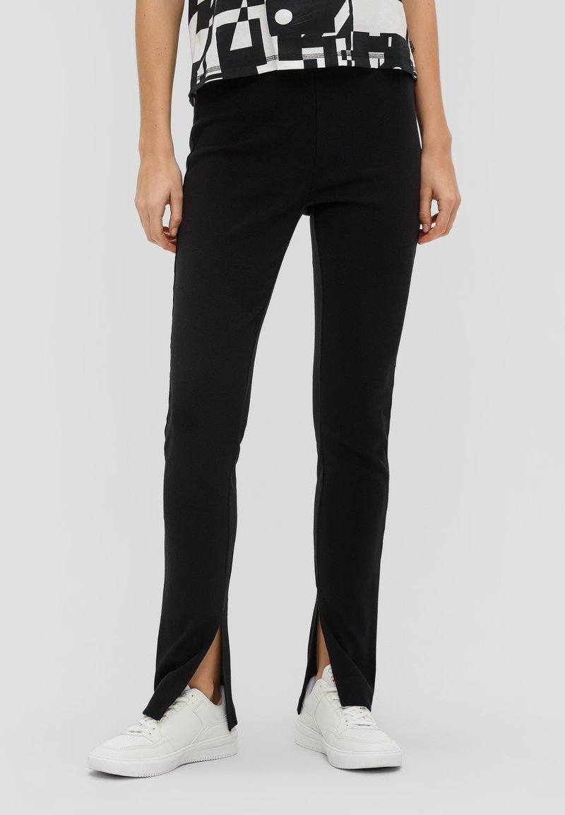 Pantalons noirs extensibles avec fentes sur les côtés, présentant une texture lisse et une coupe slim. Portés avec des baskets blanches et un haut à motif monochrome.