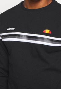 Sweat-shirt noir avec des rayures horizontales blanches et argentées, et un petit logo rouge, jaune et noir. Fabriqué en tissu doux et texturé.