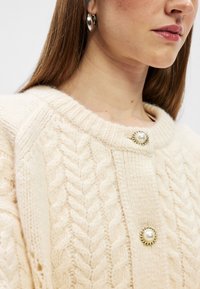 Cárdigan de punto trenzado en color crema suave, con un escote redondo y detalles decorativos de botones perlados. La textura es esponjosa y acogedora.