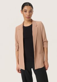 Světlý béžový blazer s volným střihem, designem na jedno tlačítko a ohrnutými rukávy, nošený přes černý pruhovaný top a černé kalhoty.