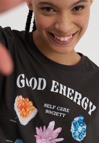 T-shirt en coton noir arborant des impressions graphiques colorées de cristaux et le texte "GOOD ENERGY SELF CARE SOCIETY" en caractères gras blancs.