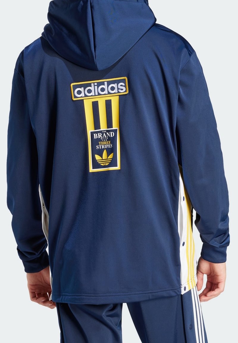 Marinblå jacka med gula detaljer och vita sidostråkar. Har Adidas-logotyp och branding på baksidan. Slät tyg, med huva, lång ärm.