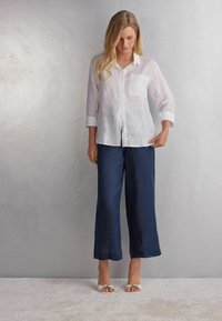 Bílá lněná košile s knoflíky a kapsou na prsou, kombinovaná s námořnickými širokými kalhotami. Model má na sobě bílé sandály. Hladká, lehká tkanina.