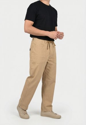 Homme portant une chemise noire à manches courtes, un pantalon beige à cordon avec de grandes poches, et des baskets décontractées beiges sur un fond uni.