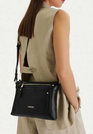 Donna che indossa un completo smanicato beige e porta una borsa a tracolla nera Lasocki con cerniera dorata e logo sul retro.