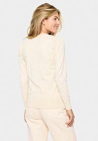 Bež pulover iz mehkega pletenega materiala, z okroglo izrezom in naborki na rokavih. Silhueta je ohlapna, z rebrastimi manšeti in robom.