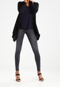 Svart öppen framsidan kavaj över en marinblå v-ringad topp, i kombination med mörkgrå skinny jeans och svarta strappy klackar. Slät textur genomgående.