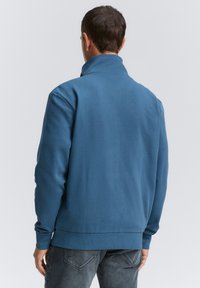 Blå sweatshirt med høj krave, ribbede manchetter og kant, lavet af blødt stof. Båret med mørke jeans, viser en ensfarvet rygvisning.