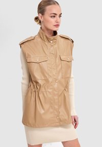 Femme portant un gilet en cuir beige sans manches avec des poches avant et des boutons-pression, par-dessus une robe crème côtelée, posant devant un fond uni.