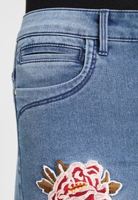 Jean de mezclilla azul con un diseño ajustado, que presenta un bolsillo lateral y una gran rosa bordada en rojo, blanco y marrón en la parte superior de la pierna.