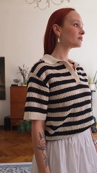 Čierny a krémový pruhovaný pletený polo sveter s krátkymi rukávmi a golierom. Nosený cez bielu sukňu, odhaľujúci kvetinové tetovanie na ruke.