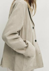 Beige mokkanahkakangas takki, jossa on rento leikkaus, suuret etunapit ja sivutaskut. Suunniteltu sileäksi rakenteeksi puhtaalla ja minimalistisella muotoilulla.