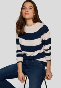 Gestreifter Pullover in Marineblau und Creme mit langen Ärmeln und Rundhalsausschnitt. Getragen zu dunkelblauen Jeans, sitzend auf einem Holzstuhl.