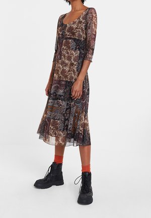Femme portant une robe patchwork fleurie mi-longue avec des manches trois-quarts, des chaussettes orange et des bottines noires à lacets.
