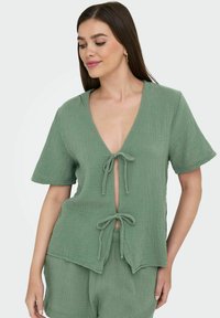 Top verde chiaro a maniche corte in un tessuto strutturato. Presenta un profondo scollo a V e chiusure a fiocco sul davanti. Fit casual e rilassato.