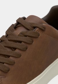 Tenisi din piele maro cu vârf rotunjit, suprafață texturată, șireturi plate maro și talpă de culoare deschisă; au un finisaj neted și detalii minime.