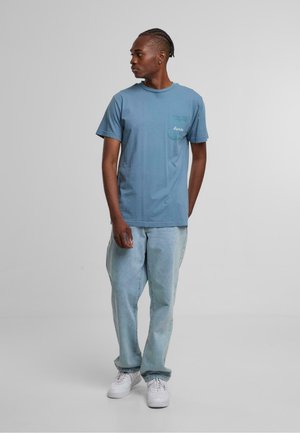 Lys blå bomull t-skjorte med en brystlomme, sammen med falmede blå denim jeans og hvite sneakers, stående mot en nøytral bakgrunn.