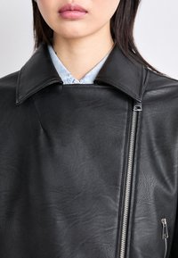 Giacca in pelle nera con un ampio colletto, chiusura con zip asimmetrica e tasca con zip. Texture liscia con un delicato dettaglio a grana.