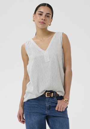 Femme aux cheveux foncés portant un top blanc sans manches à col en V avec de fines rayures, un jean bleu, des boucles d'oreilles créoles dorées et un bracelet en or.