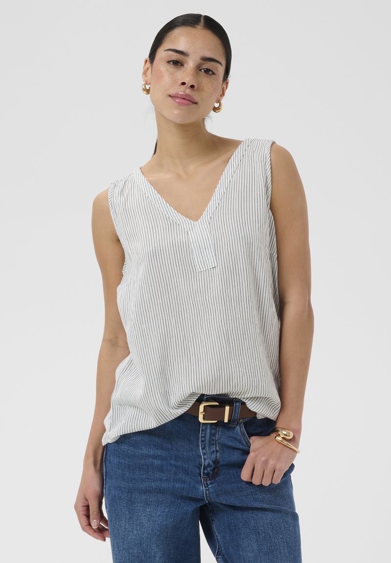 Femme aux cheveux foncés portant un top blanc sans manches à col en V avec de fines rayures, un jean bleu, des boucles d'oreilles créoles dorées et un bracelet en or.