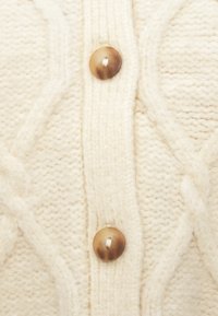 Πλεκτό ύφασμα χρώματος κρέμα με σχέδια καλωδίων και δύο στρογγυλά καφέ κουμπιά ραμμένα κάθετα στο κέντρο.