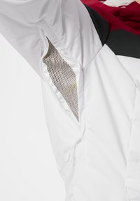 Chaqueta blanca con un bolsillo interior de malla. El diseño incluye un bloque de color rojo y negro en la parte superior y cordones elásticos.