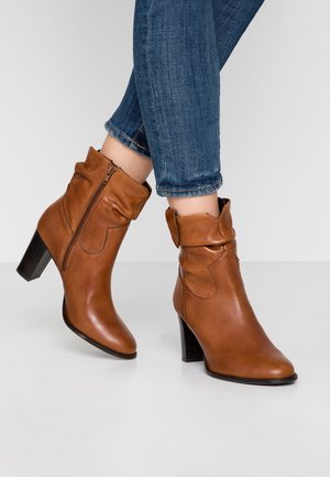 Braune Leder-Stiefeletten mit schlabberigem Design, seitlichem Reißverschluss und gestapeltem Absatz, kombiniert mit blauen Jeans. Glatte Textur und schlanke Silhouette.