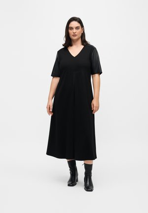 Femme aux cheveux foncés mi-longs, vêtue d'une robe midi noire à col en V, manches courtes et bottines noires, debout sur un fond blanc.