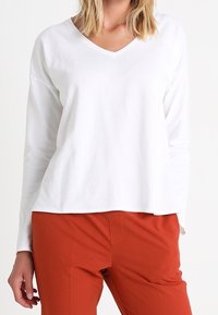 White long-sleeve V-neck top with a relaxed fit, paired with high-waisted orange pants. Smooth fabric, minimalistic design.