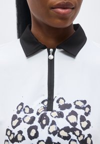 Κοντινό πλάνο ενός ατόμου που φοράει λευκό πουκάμισο με μαύρο γιακά, φερμουάρ και ασπρόμαυρο-μπεζ λεοπάρ μοτίβο διακοσμημένο με μικρές στρας.