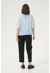 Jasnoniebieska prześwitująca bluzka z krótkimi rękawami, falbankowym kołnierzykiem i wykończeniem. W połączeniu z czarnymi zwężanymi spodniami i masywnymi czarnymi butami.