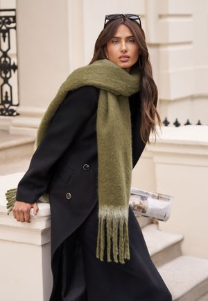 Žena s dugom smeđom kosom, odjevena u crni kaput i preveliki maslinasto zeleni šal, naslonjena na bijelu kamenu ogradu na otvorenom.