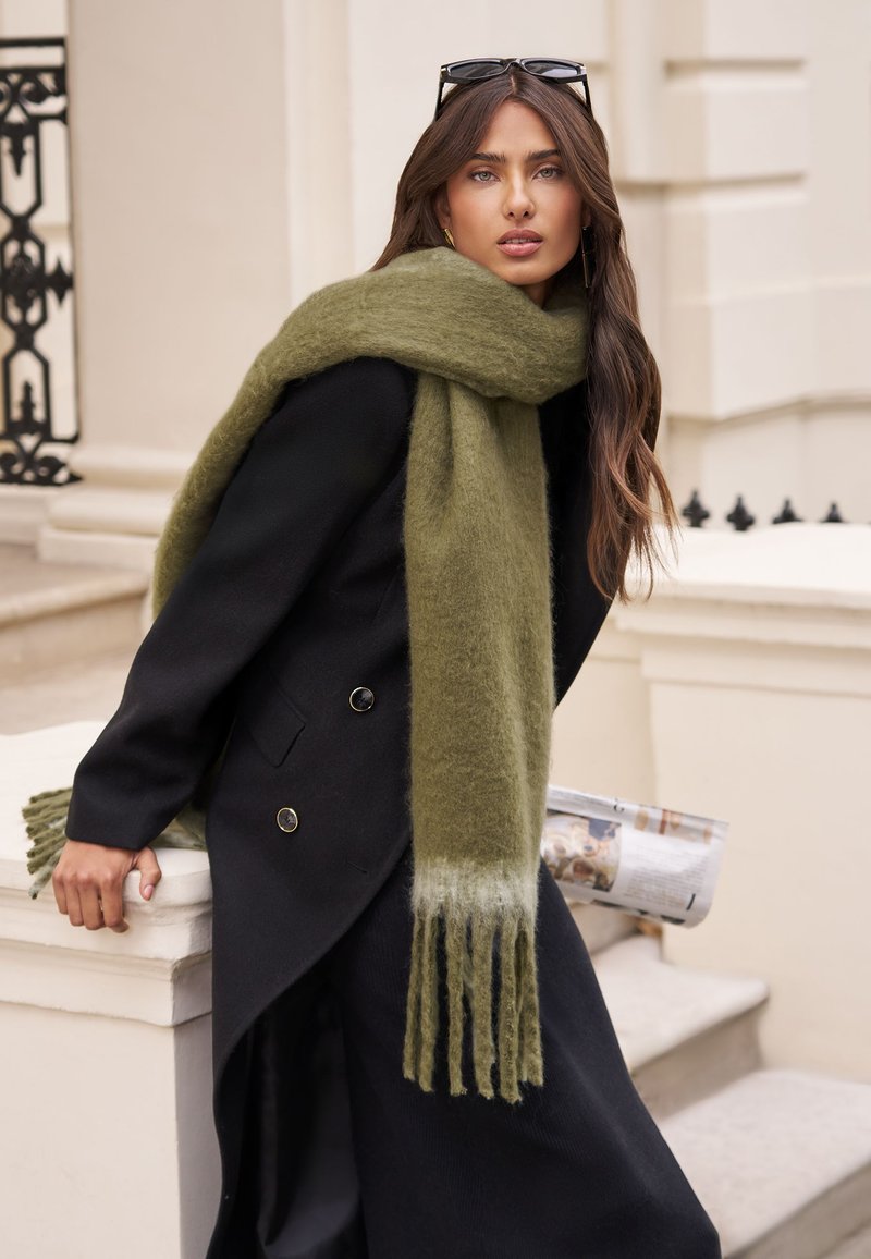 Ženska z dolgimi rjavimi lasmi, oblečena v črni plašč in prevelik olivno zelen šal, ki se naslanja na belo kamnito ograjo na prostem.