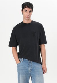 Schwarzes T-Shirt mit kurzen Ärmeln aus Baumwolle, mit einem tonal gestickten Logo auf der Brust, kombiniert mit hellblauen Jeans.