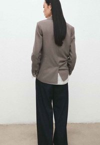 Geruite blazer met een getailleerde pasvorm en een gespleten achterkant, gecombineerd met losse donkere broek. Het materiaal lijkt textuurrijk en licht van gewicht.