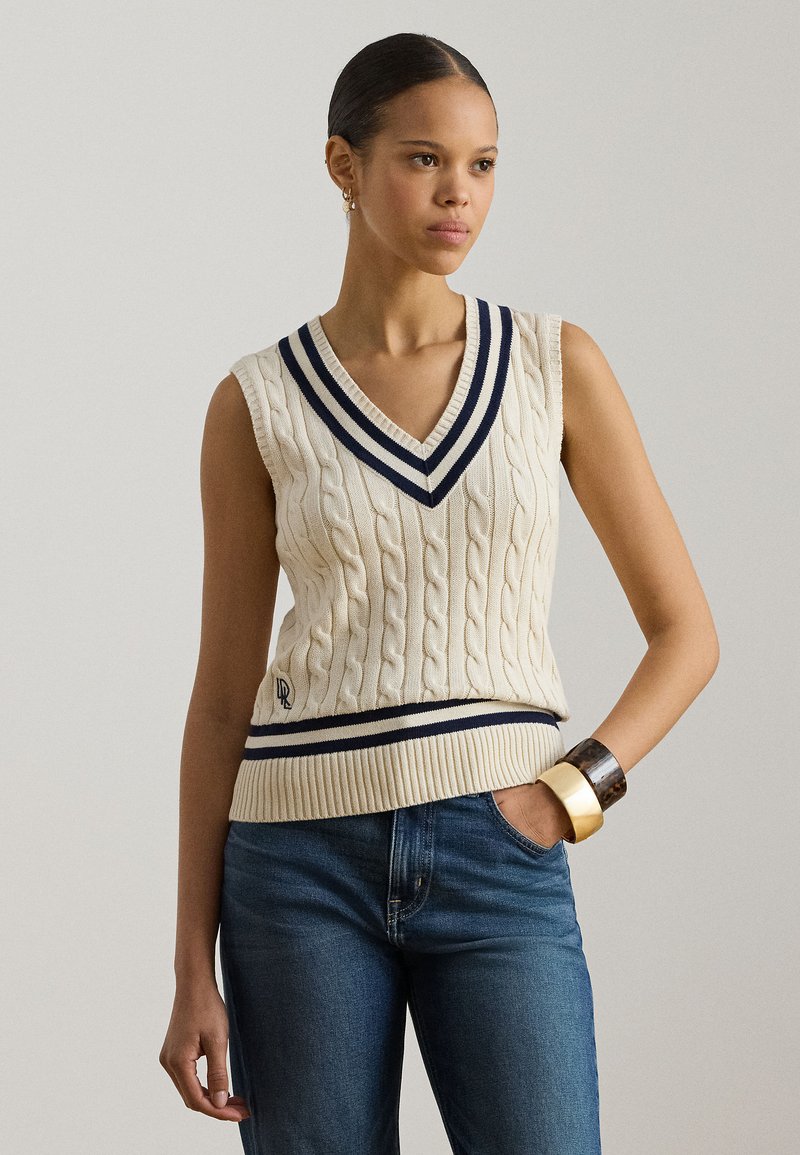 Κρεμ γιλέκο με πλέξη καλωδίου, βαθύ V-λαιμό, ναυτικές ρίγες στον λαιμό και στο τελείωμα, και ριμπ ζώνη στη μέση. Φορεμένο πάνω από μπλε τζιν.