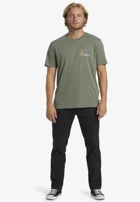 Olive green cotton t-shirt with a small multicolor "Billabong" logo on the chest. Paired with black pants and black canvas shoes.