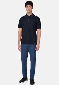 Marineblaues Polohemd mit kurzen Ärmeln und Reißverschluss am Ausschnitt, kombiniert mit blauen Sporthosen und schwarzen Turnschuhen auf neutralem Hintergrund.