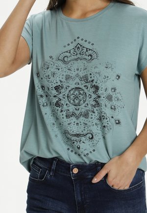 Femme portant un t-shirt bleu clair avec un imprimé mandala noir complexe, associé à un jean denim bleu foncé, main dans la poche.