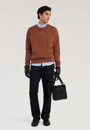 Mladý muž v hnědé mikině, modré košili, černých kalhotách a černých rukavicích, držící černou tašku přes rameno, dívající se stranou na jednobarevném pozadí.