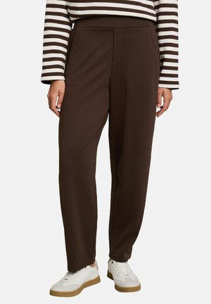 Persoon draagt donkerbruine, rechte broek met zijzakken, een donkerbruine en wit gestreepte top met lange mouwen, en witte schoenen met rubberzolen.