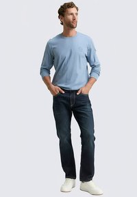 Hellblaues Langarmshirt mit rundem Ausschnitt und Logodetail, kombiniert mit dunklen Jeans und weißen Sneakers.