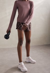 Světle fialové tričko s dlouhým rukávem, vzorované hnědé šortky, bílé kotníkové ponožky a růžové sportovní boty. Drží černý kettlebell.