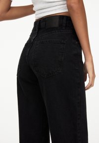 Jean en denim noir à taille haute, coupe droite, deux poches arrière et une étiquette en cuir noir à la ceinture.